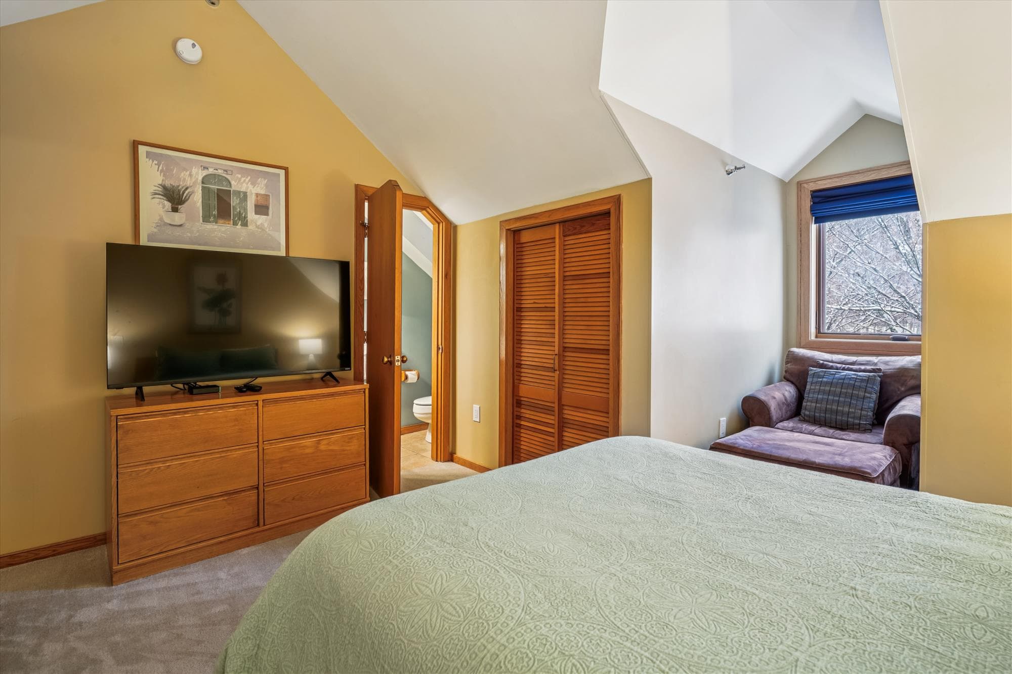 Comfortable queen bedroom with a flat-screen TV, dresser storage, and a sitting chair by the window.