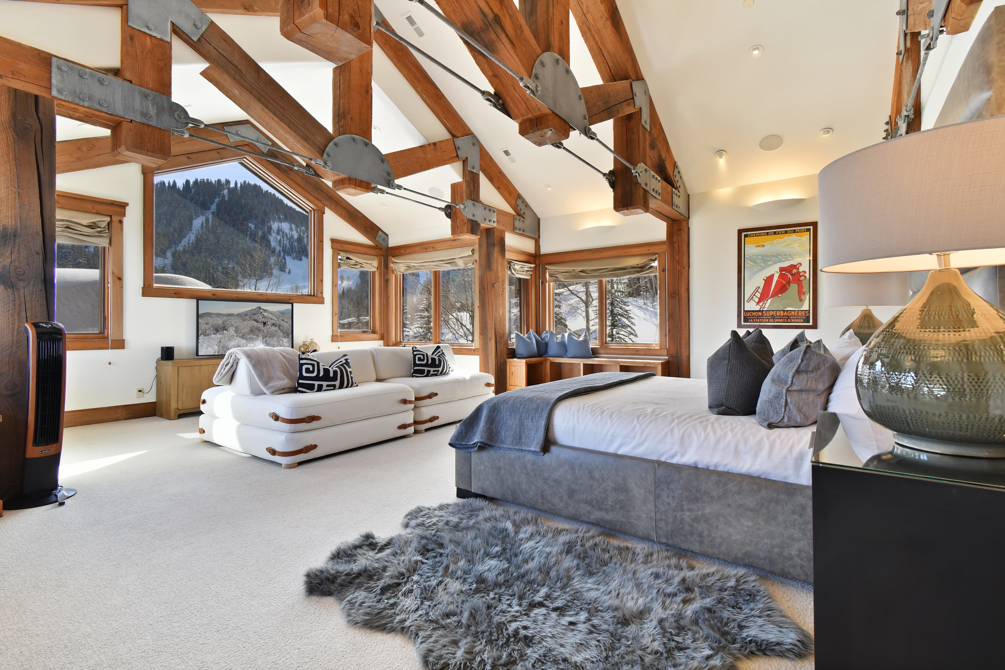 Large windows sit in front of the bed of the primary bedroom, with a large couch in front of an entertainment center and TV.