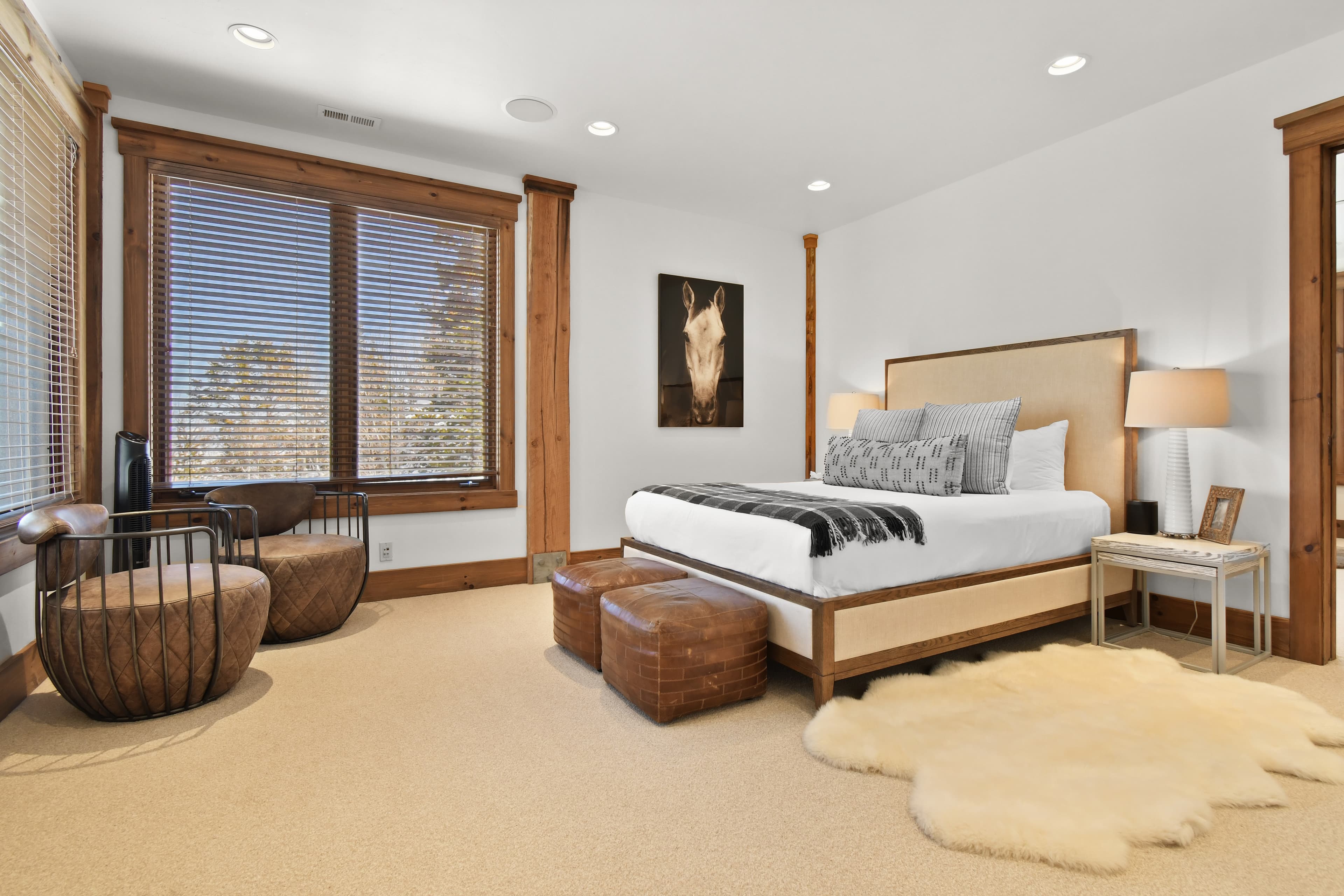 A white fur rug is set beside a large bed, with two leather ottomans at the foot of it.
